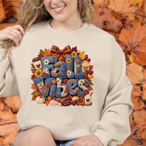 May include: A beige crewneck sweatshirt featuring a graphic print with the text Fall Vibes in blue denim-textured letters. The design is surrounded by autumn-themed elements including orange leaves, sunflowers, pumpkins, a candle, and coffee cups.