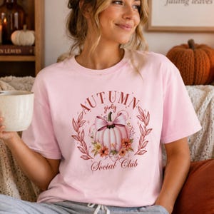 May include: A light pink t-shirt featuring a graphic of a pale pink pumpkin adorned with a large bow and surrounded by floral accents. The text reads Autumn girly Social Club in a decorative font, framed by two curved leafy branches.