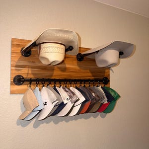 May include: A wooden hat rack with two cowboy hats and a row of baseball caps. The rack is made of a wooden board with black metal pipe fittings. The hats are various colors, including beige, white, and navy blue.