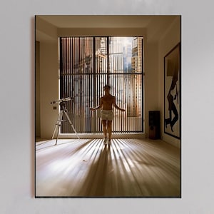 Könnte beinhalten: Gerahmtes Foto einer Person in einem Raum mit einem Teleskop und einem großen Fenster. Die Person trägt weiße Shorts und steht vor dem Fenster. Der Raum hat Holzböden und vertikale Jalousien.