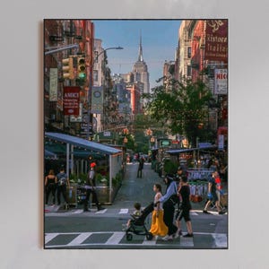 May include: A vertical photograph of a New York City street scene featuring a crosswalk with pedestrians and a stroller. The Empire State Building is visible in the distance between brick buildings. Red signs for Amici and Ristorante are present.