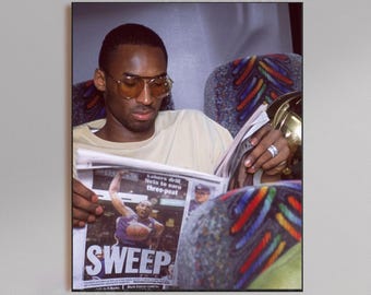 May include: A framed print featuring a man wearing glasses reading a newspaper with the headline "SWEEP". The newspaper shows a basketball player in action. The man is wearing a light-colored shirt.