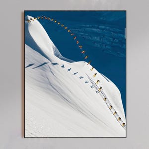 May include: A framed print featuring a sequence of skiers descending a snow-covered mountain. The skiers are wearing yellow and orange outfits, creating a contrast against the white snow and blue sky. The image captures the skiers' movements down the slope.