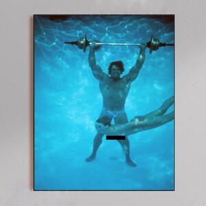 May include: A framed photograph depicts two people underwater in a swimming pool. One person is lifting a barbell overhead, while the other is swimming below. The water is a bright blue, and the image has a vintage aesthetic.