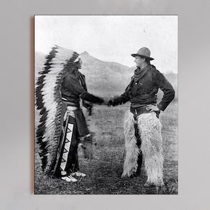 Puede incluir: Fotografía en blanco y negro de dos personas dándose la mano. Una persona lleva un tocado y ropa tradicional nativa americana. La otra persona lleva sombrero, bufanda, chaqueta y chaparreras con flecos. El fondo muestra una cordillera.