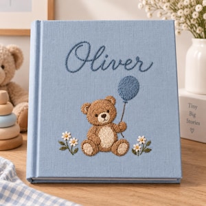 Può includere: Un album fotografico rivestito in tessuto azzurro con dettagli ricamati sulla copertina. Il nome Oliver è cucito in filo blu scuro sopra un orsacchiotto marrone che tiene un palloncino blu, affiancato da piccoli fiori di margherita bianchi.