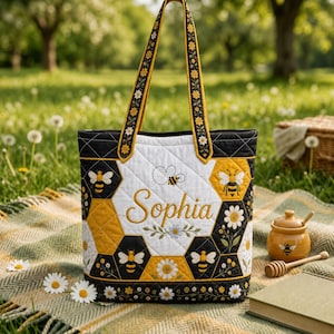 May include: A quilted tote bag featuring a honeycomb pattern in black, white, and yellow. The design includes embroidered bees, daisies, and the name Sophia in cursive script. It has two long shoulder straps with a matching floral pattern.