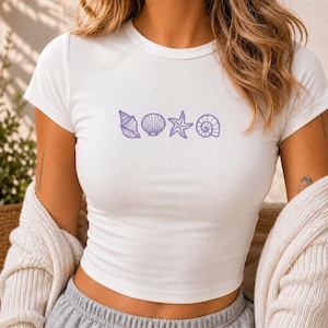 May include: White crop top with a purple seashell design. The design features a conch shell, scallop shell, starfish, and spiral shell. The shirt has short sleeves and a crew neck.