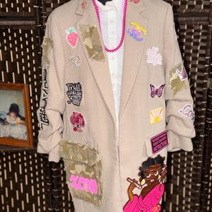 May include: A beige blazer adorned with various colorful patches, including a strawberry, a butterfly, and a cartoon character in a pink robe. The blazer features camouflage fabric accents on the pockets and sleeves, paired with a white shirt and a bright pink beaded necklace.