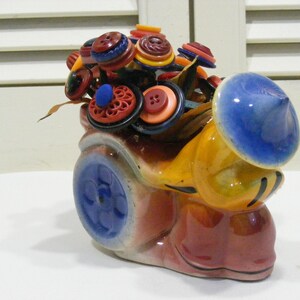 May include: A whimsical ceramic snail-shaped vase, painted in shades of red, yellow, and blue. The snail's shell is filled with colorful buttons, resembling a bouquet of flowers. The vase has a blue wheel and a blue hat.