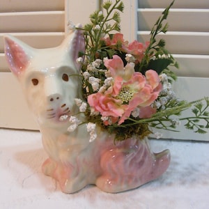 May include: A white ceramic dog planter with pink accents, holding a bouquet of pink flowers and greenery.