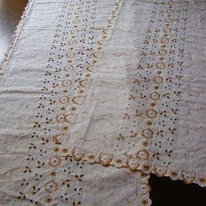 May include: A white, rectangular tablecloth with intricate cutwork and embroidered floral and leaf patterns. The edges are scalloped and trimmed with a delicate, embroidered border. The design features repeating floral motifs and openwork details.