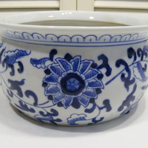 May include: A white ceramic planter with a blue floral design. The planter has a rounded shape with a slightly flared rim. The blue floral pattern includes a large flower in the center and smaller floral and leaf motifs around the sides.