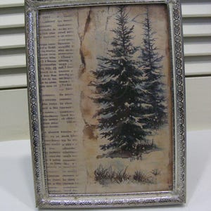 May include: A rectangular silver-colored picture frame with a winter scene. The artwork features two evergreen trees covered in snow, set against a backdrop of aged paper with text. The frame has a detailed, ornate border, adding a vintage aesthetic.