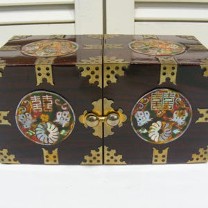 May include: A dark brown wooden box with brass accents and decorative circular inlays. The box features a double-door design with a brass clasp and corner brackets. The inlays display colorful floral and butterfly motifs.