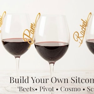 Può includere: Tre calici di vino rosso, ognuno con un'etichetta dorata con i nomi "Smelly Cat", "Pam" e "Rachel". Il testo "Build Your Own Sitcom Set" è sotto, con le parole "Beets, Pivot, Cosmo, Schrute".