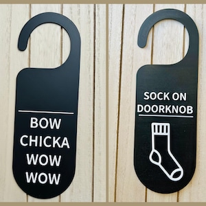 Pode incluir: Dois cabides de porta pretos com uma parte superior curva. Um diz "BOW CHICKA WOW WOW" em texto branco. O outro diz "SOCK ON DOOR KNOB" com um desenho de meia branca.