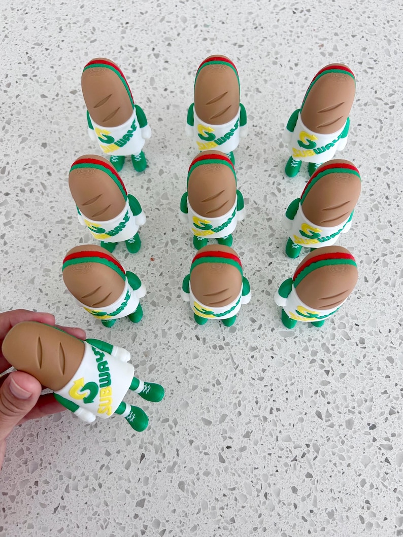 May include: A collection of ten small Subway-themed figurines, each featuring a tan bread-shaped head with a red stripe, a white shirt with the green and yellow Subway logo, and green arms and legs. The figures are arranged in a grid pattern.