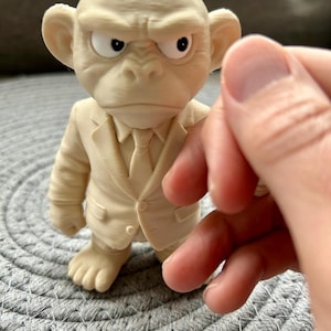 May include: A cream-coloured 3D printed figurine of a monkey wearing a suit and tie. The figure has a stern facial expression with furrowed brows and white eyes with black pupils. The texture shows fine horizontal lines from the printing process.