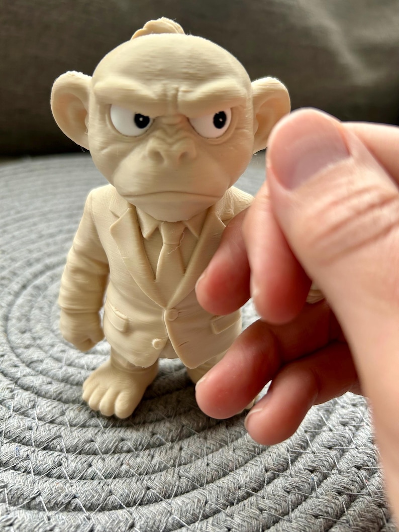 May include: A cream-coloured 3D printed figurine of a monkey wearing a suit and tie. The figure has a stern facial expression with furrowed brows and white eyes with black pupils. The texture shows fine horizontal lines from the printing process.