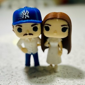 May include: Two small stylised figurines standing side by side. The left figure wears a blue baseball cap with a white NY logo, a white collared shirt, and blue trousers. The right figure has long brown hair and wears a simple white dress.
