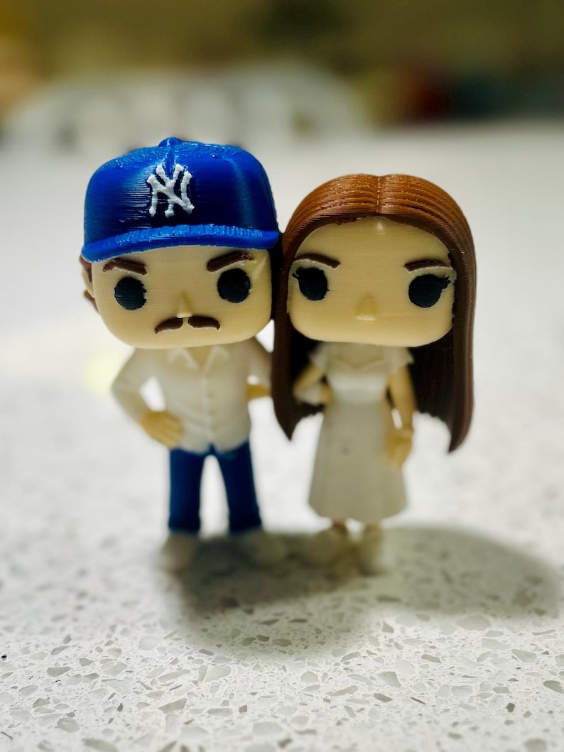 May include: Two small stylised figurines standing side by side. The left figure wears a blue baseball cap with a white NY logo, a white collared shirt, and blue trousers. The right figure has long brown hair and wears a simple white dress.