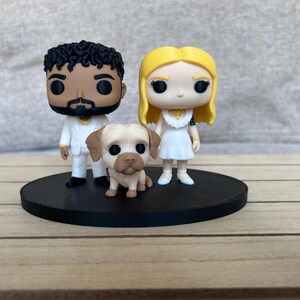May include: Two stylised vinyl figures and a small dog figurine standing on a circular black base. One figure has dark curly hair and a white suit, the other has long blonde hair and a white dress. The dog is tan with brown ears and a dark muzzle.