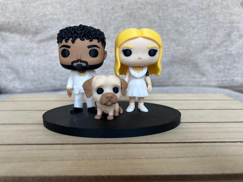 May include: Two stylised vinyl figures and a small dog figurine standing on a circular black base. One figure has dark curly hair and a white suit, the other has long blonde hair and a white dress. The dog is tan with brown ears and a dark muzzle.