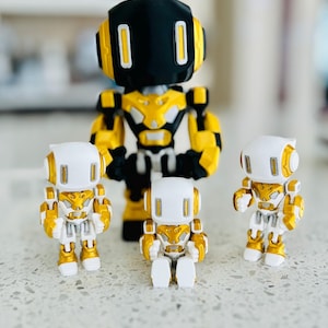 May include: A collection of four robot figurines. One large robot features a black head with white rectangular eyes, a gold and black torso, and gold arms. Three smaller robots feature white heads with gold accents, white torsos, and gold limbs.