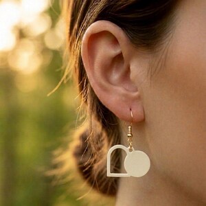 May include: Cream-colored geometric earrings with a gold-colored hook. The earrings feature a square shape with a rounded top and a circle shape. The earrings are hanging from an ear.