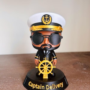 May include: A figurine of a captain wearing a white and black hat with a gold anchor emblem, black sunglasses, and a black uniform with gold accents. The figure holds a gold ship wheel and stands on a black base with the text Captain Delivery.