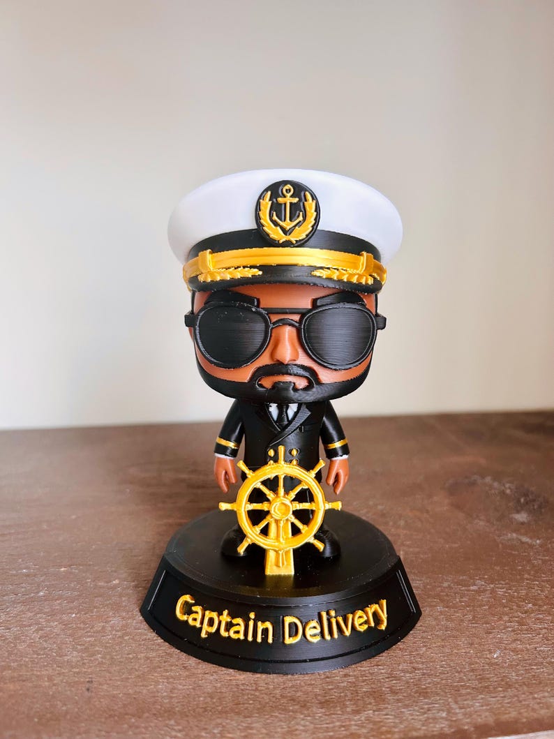 May include: A figurine of a captain wearing a white and black hat with a gold anchor emblem, black sunglasses, and a black uniform with gold accents. The figure holds a gold ship wheel and stands on a black base with the text Captain Delivery.