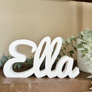Peut inclure: Un panneau en bois blanc avec le nom "Ella" écrit en cursive. Le panneau est posé sur une étagère en bois avec de la verdure et un pot blanc en arrière-plan. Le panneau est un objet décoratif.