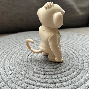 May include: A small, light beige, 3D-printed figurine of a monkey wearing a collared jacket. The monkey is depicted in profile, showing a curved tail, rounded ears, a textured head, and a smooth, solid-coloured surface throughout the entire figure.