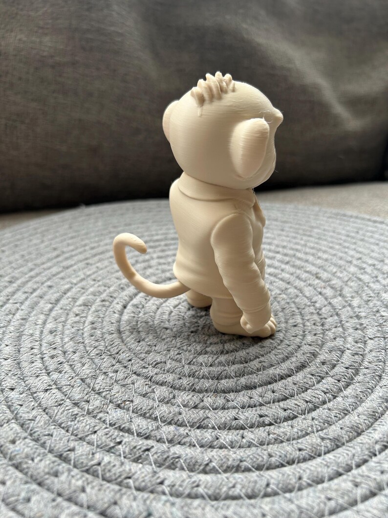 May include: A small, light beige, 3D-printed figurine of a monkey wearing a collared jacket. The monkey is depicted in profile, showing a curved tail, rounded ears, a textured head, and a smooth, solid-coloured surface throughout the entire figure.