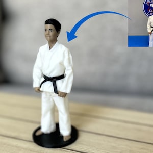 May include: A 3D printed figurine of a person wearing a white martial arts uniform with a black belt, standing on a circular black base. A blue arrow points from a small inset photograph of a person in the same uniform to the figurine.