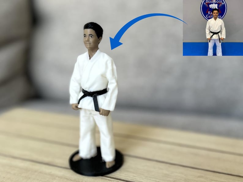 May include: A 3D printed figurine of a person wearing a white martial arts uniform with a black belt, standing on a circular black base. A blue arrow points from a small inset photograph of a person in the same uniform to the figurine.