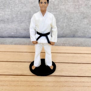 May include: A figurine of a person with dark hair wearing a white martial arts uniform, known as a gi, with a black belt tied around the waist. The figure is standing in a neutral pose on a circular black base, with bare feet visible at the bottom.