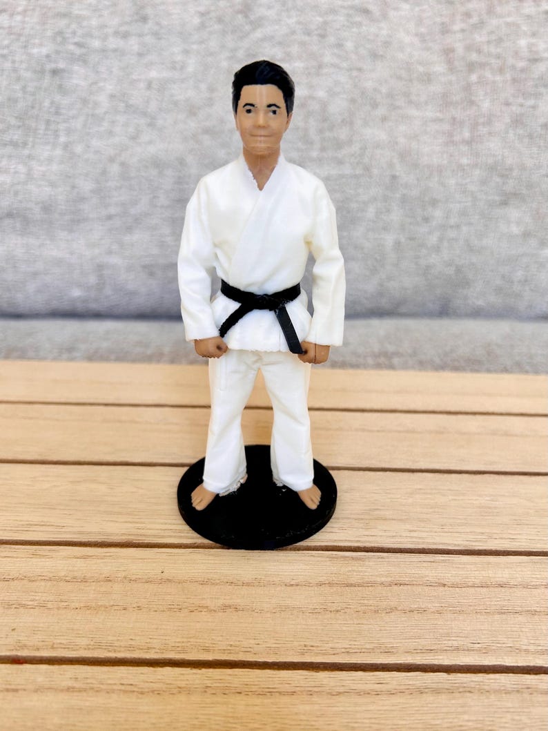 May include: A figurine of a person with dark hair wearing a white martial arts uniform, known as a gi, with a black belt tied around the waist. The figure is standing in a neutral pose on a circular black base, with bare feet visible at the bottom.