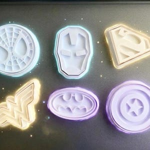 Puede incluir: Un juego de seis cortadores de galletas de plástico blanco con varios emblemas de superhéroes, incluyendo una máscara de telaraña, una máscara robótica, un logotipo de escudo triangular, un emblema alado, un símbolo de murciélago y un escudo circular con una estrella central.