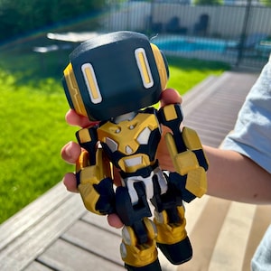 May include: A small robot figurine featuring a black head with two vertical white rectangular eyes, a gold and black chest plate, and articulated gold and black limbs. The robot has a blocky, mechanical design with visible layered textures.