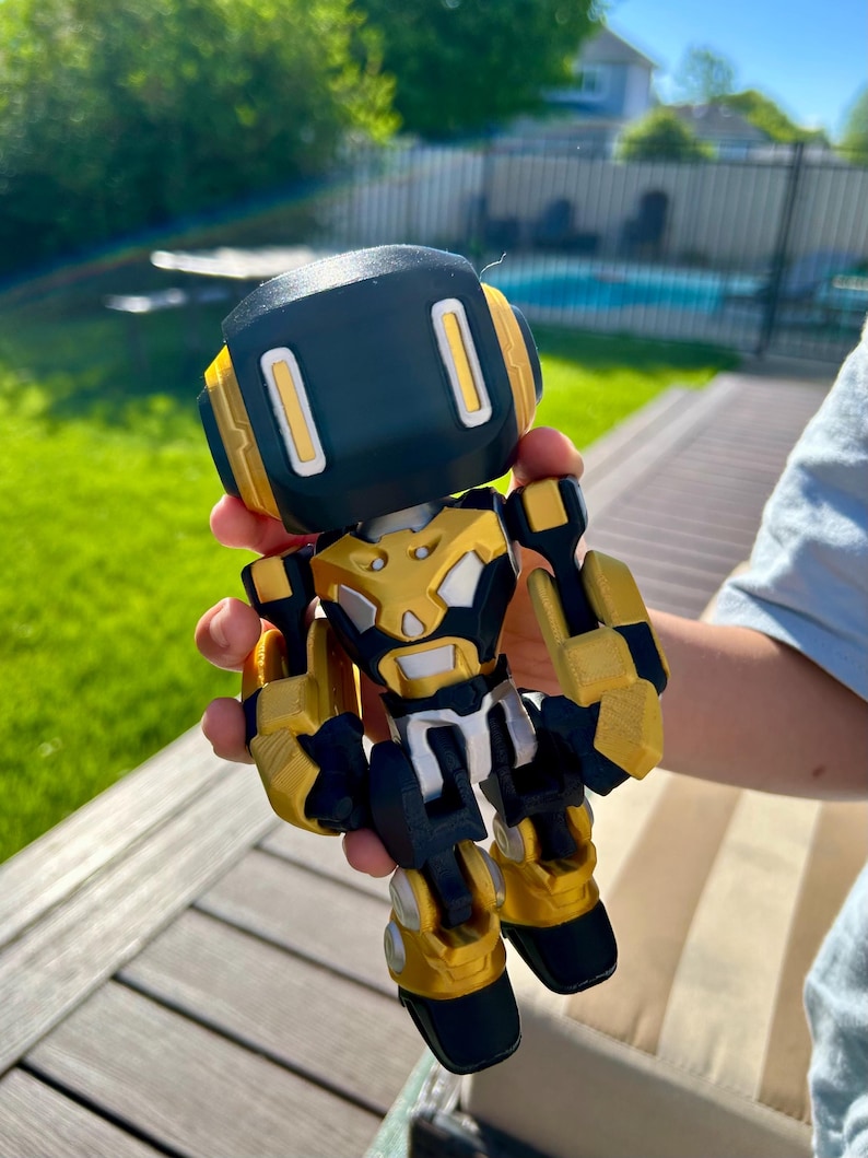 May include: A small robot figurine featuring a black head with two vertical white rectangular eyes, a gold and black chest plate, and articulated gold and black limbs. The robot has a blocky, mechanical design with visible layered textures.
