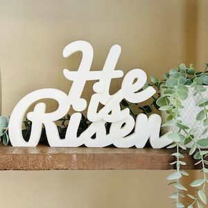 Puede incluir: Un letrero de madera blanco con las palabras "He is Risen" en una fuente cursiva. El letrero se muestra en un estante marrón, con vegetación y una maceta blanca en el fondo. El letrero es un artículo decorativo.