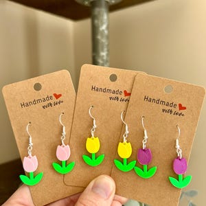 Könnte beinhalten: Handgefertigte Tulpenohrringe in Rosa, Gelb und Lila. Jedes Paar hat ein farbenfrohes Tulpendesign mit grünen Blättern und silberfarbenen Haken. Die Ohrringe werden auf braunen Karten mit dem Text "Handmade with love" präsentiert.