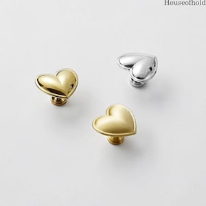 May include: Three heart-shaped cabinet knobs in gold and silver. The knobs have a smooth, polished finish and are mounted on a small base. The gold knobs have a warm, metallic sheen, while the silver knob is bright and reflective. The knobs are decorative hardware.