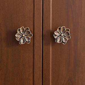 Peut inclure: Deux poignées de porte en forme de fleur de couleur bronze, montées sur une porte d'armoire en bois brun foncé. Les poignées présentent des motifs de pétales détaillés et un accent central circulaire. Le texte "luxury hardware" est visible en haut à gauche et "HouseofholdZ" en haut à droite.