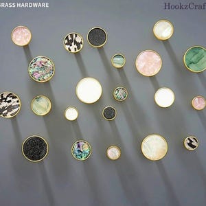 May include: A selection of decorative brass hardware knobs in various sizes and designs. The knobs feature mother-of-pearl, speckled black, and patterned surfaces, all set within gold-toned brass frames. The background is a solid grey.