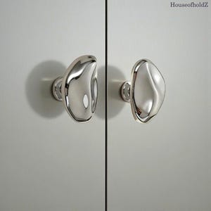 May include: Two polished silver cabinet knobs with an organic, flowing design. The knobs are mounted on a light grey cabinet door, casting a soft shadow. The knobs have a unique, irregular shape, adding a modern touch to the cabinet.
