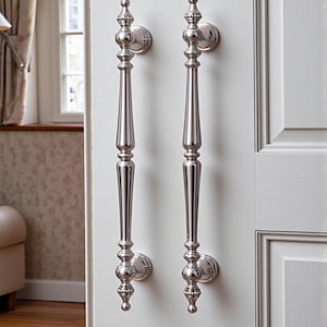May include: Two polished silver door pulls with ornate detailing are mounted on a white door. The pulls have a long, slender design with decorative finials at the top and bottom. The door is part of a white door frame.