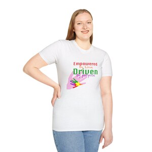 May include: A white t-shirt with the text "Empowered by Love, Driven by Purpose" in red and green. The shirt features a graphic of a hand with colorful fingers. The person is wearing blue jeans.